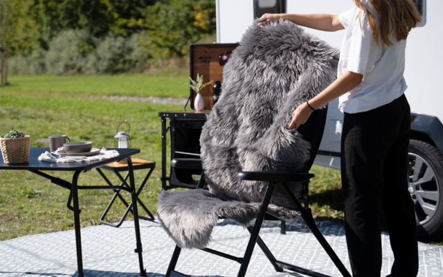 Teil einer Person neben einem Camping-Klappstuhl mit grauem Fellüberwurf, kleiner Klapptisch mit Geschirr, offene Wohnmobiltür, Gras und Bäume