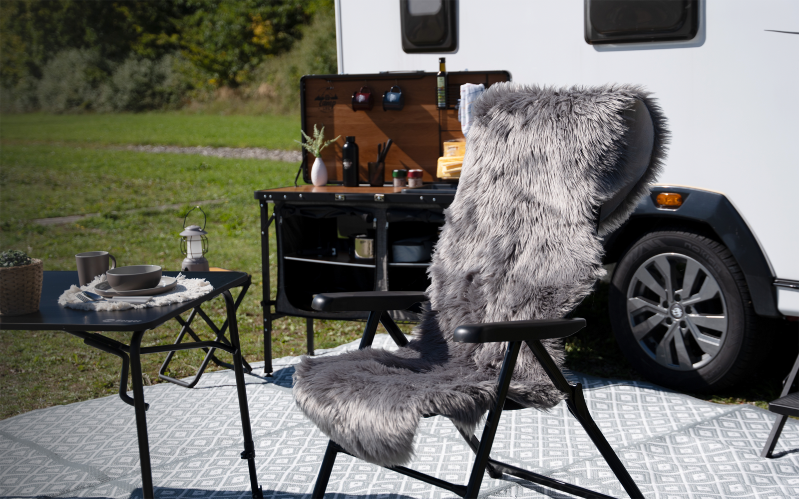 Klappstuhl mit grauem Kunstfellbezug vor einem Wohnwagen, kleiner Tisch mit Geschirr und tragbarer Camping-Küche, Rasen im Hintergrund