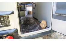 Offener Fahrzeugeingang mit Blick ins Innere: dunkler Fellteppich auf Holzfußboden, Rattanlaterne an der Türschwelle, graue Sitzbank rechts, Schrank mit Schubladen, obere Liege mit blauem Bezug, Rattankorb, Kiesboden außen und Rücklicht des Fahrzeugs