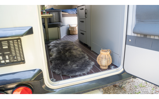 Offener Fahrzeugeingang mit Blick ins Innere: dunkler Fellteppich auf Holzfußboden, Rattanlaterne an der Türschwelle, graue Sitzbank rechts, Schrank mit Schubladen, obere Liege mit blauem Bezug, Rattankorb, Kiesboden außen und Rücklicht des Fahrzeugs