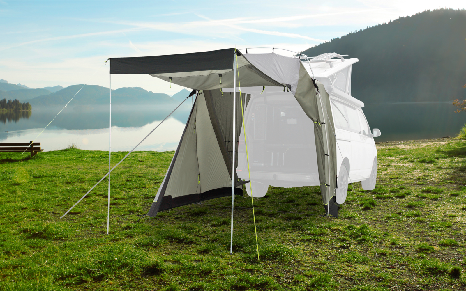 White camping tent with open entrance stands on a green meadow by a lake with mountains in the background
