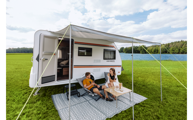 Wohnwagen mit ausgefahrener Markise auf einer Wiese; zwei Personen sitzen auf Campingstühlen an einem kleinen Tisch auf einem Teppich unter der Markise; offene Tür; See und Wald im Hintergrund; bewölkter Himmel