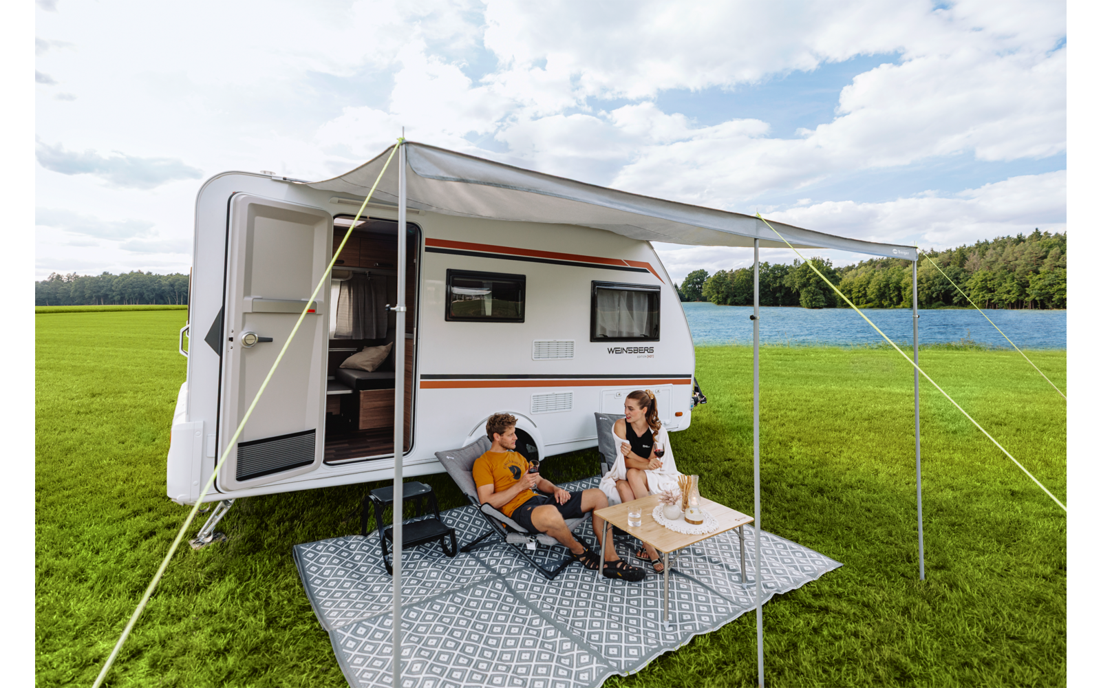 Wohnwagen mit ausgefahrener Markise auf einer Wiese; zwei Personen sitzen auf Campingstühlen an einem kleinen Tisch auf einem Teppich unter der Markise; offene Tür; See und Wald im Hintergrund; bewölkter Himmel