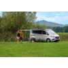 Zwei Personen stehen nebeneinander vor einem geparkten Wohnmobil auf einer Wiese; Markise ausgefahren, zwei Campingstühle und ein kleiner Tisch neben dem Fahrzeug, Bäume und Hügel im Hintergrund, bewölkter Himmel