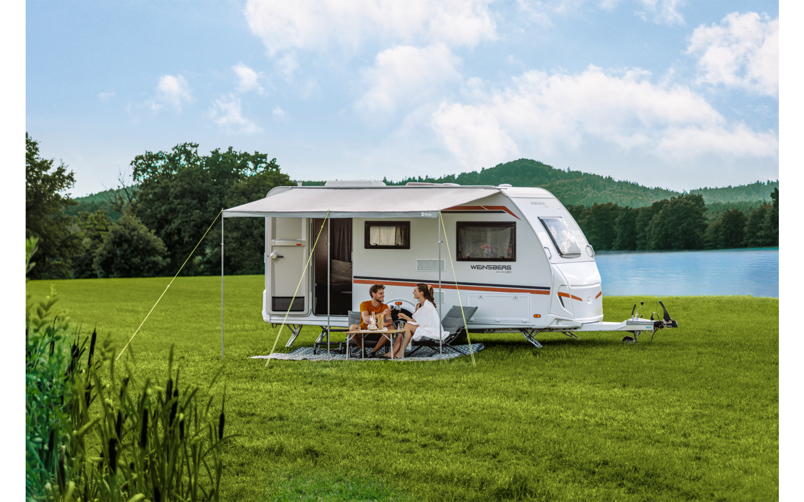 Weißer Wohnwagen mit ausgefahrenem Sonnendach auf grüner Wiese am See, drei Personen sitzen an einem Campingtisch unter dem Dach