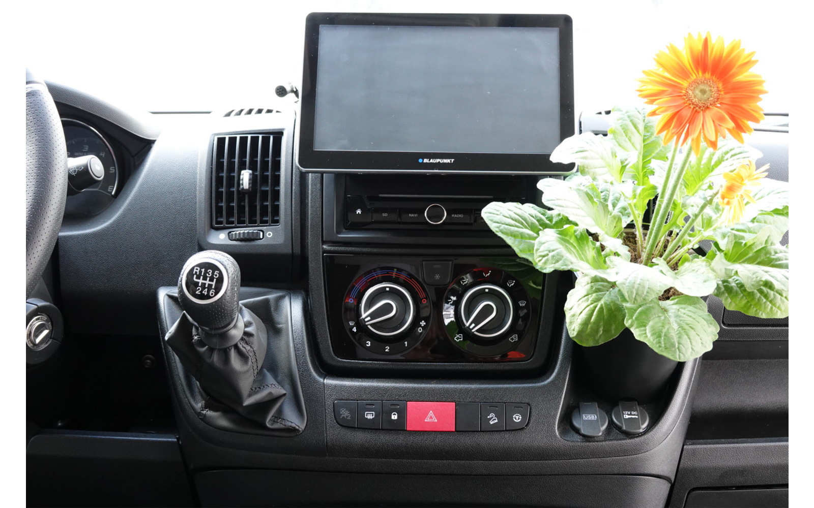 Blick auf die Mittelkonsole eines Autos mit einem Bildschirm, Klimabedienelementen und einem kleinen Blumenstrauß