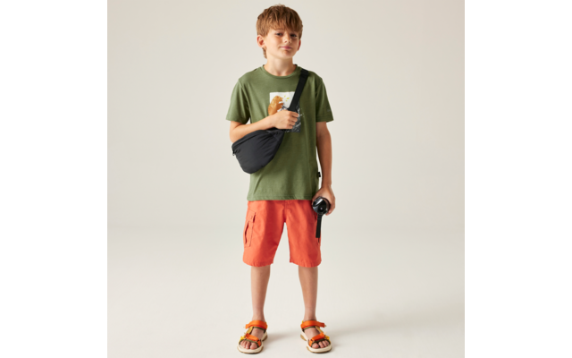 Ein Junge steht frontal vor hellem Hintergrund, trägt grünes T‑Shirt mit Motiv, orange Shorts, Sandalen und eine schwarze Umhängetasche; er hält ein kleines schwarzes Objekt mit Handschlaufe in der rechten Hand