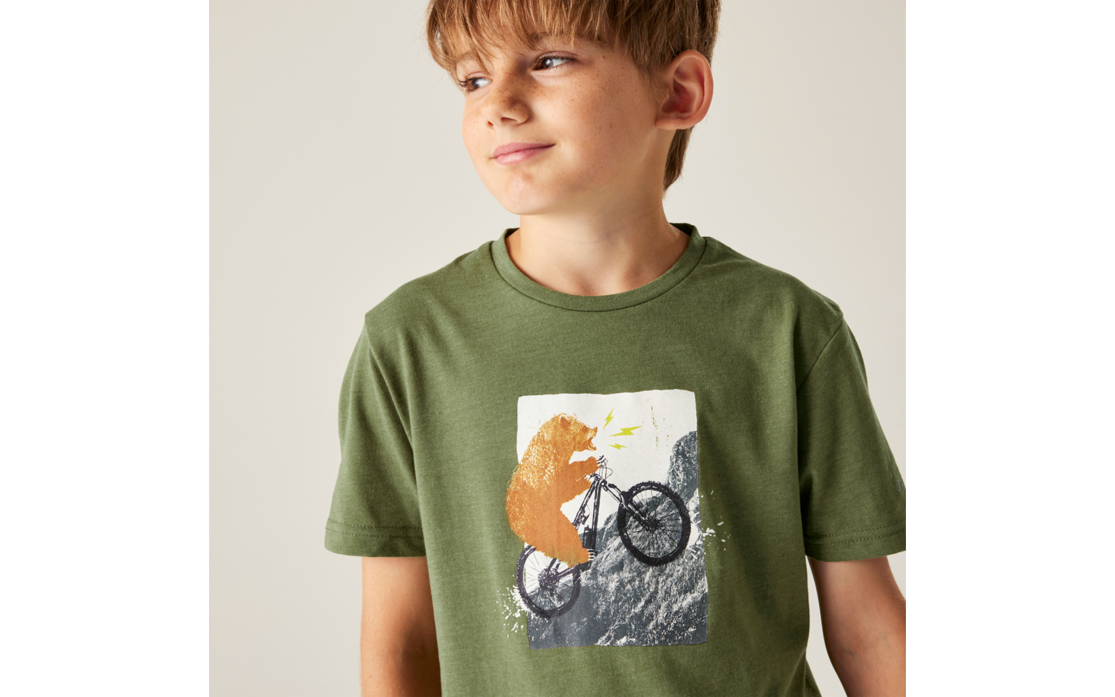 Oberkörper eines Jungen in grünem T‑Shirt, Blick nach rechts; T‑Shirt-Aufdruck: Bär auf Fahrrad auf Felsen