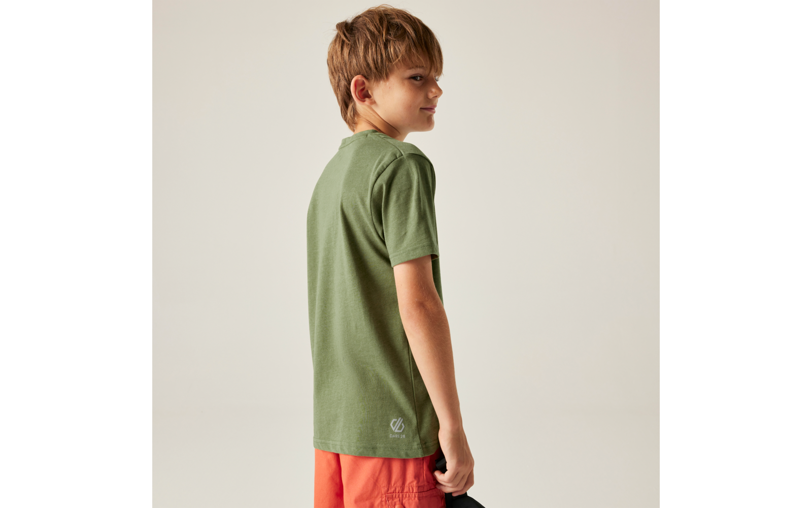 Junge, seitlich und leicht von hinten gesehen, trägt grünes T-Shirt und orange Shorts, hält eine schwarze Kappe, heller Hintergrund