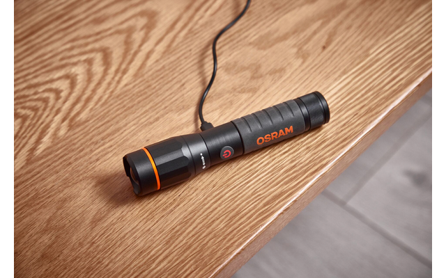 Schwarze Taschenlampe mit orangefarbenen Akzenten und 'OSRAM'-Aufschrift, angeschlossenes Kabel, liegt auf einer Holzfläche; rechts Fliesenboden sichtbar