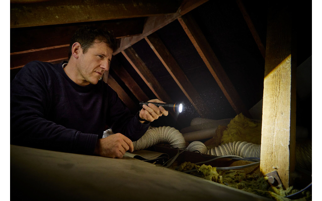 Person hält Taschenlampe in einem Dachboden mit sichtbaren Dachbalken, flexiblen Lüftungsrohren und Dämmmaterial