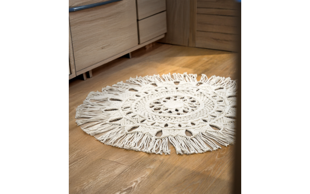 Runder, weißer Häkelteppich auf Holzboden vor einem Möbelstück aus Holz