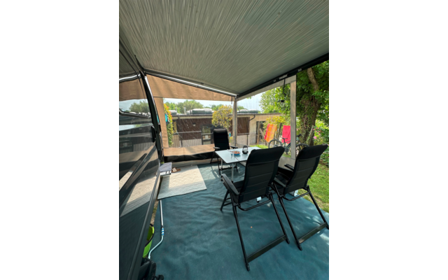 Espace assis couvert sous une marquise avec deux fauteuils pliants noirs, petite table avec verres et objets, tapis de sol ; à gauche une paroi de caravane, en arrière-plan une palissade en bois, un arbre et une chaise de camping rouge