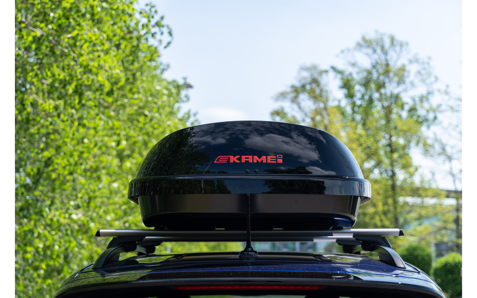 Box da tetto nero con scritta rossa KRAMER montato su portapacchi auto, alberi e cielo sullo sfondo