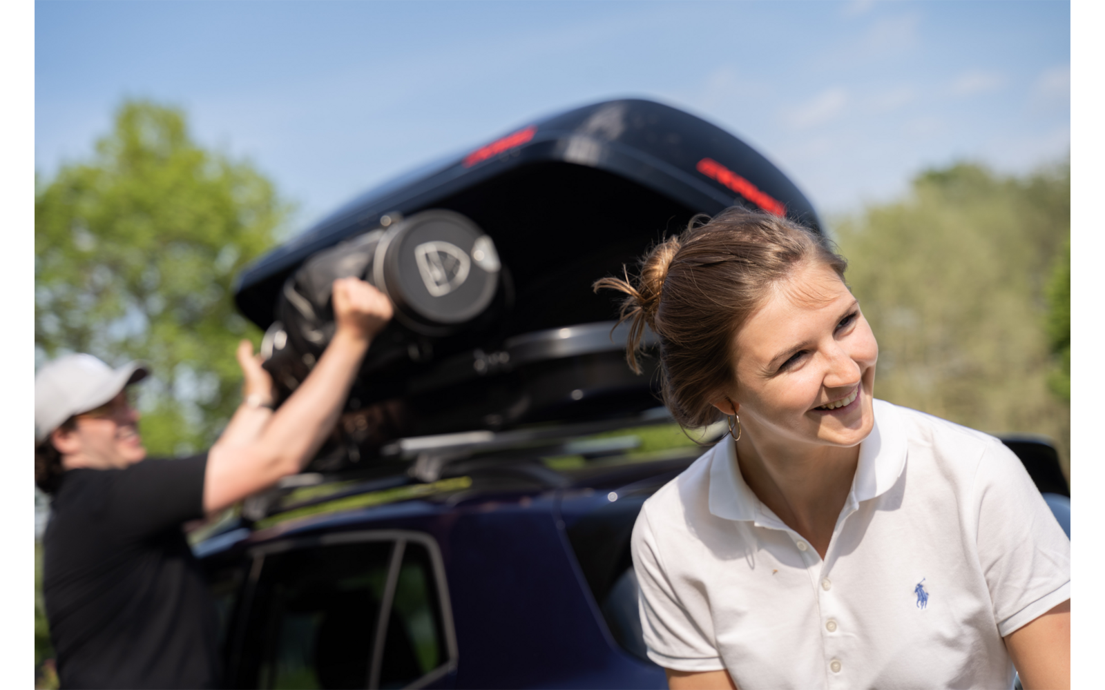Donna con capelli raccolti e polo bianca sorride davanti a un'auto; sullo sfondo, un uomo con cappellino fissa un baule da tetto nero