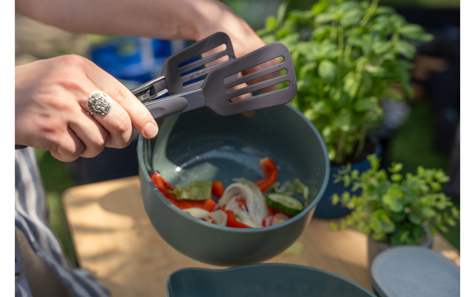 Twee handen met een ring houden twee grijze spatels en scheppen gesneden groenten (rode paprika, uienringen, groene stukjes) in een turkooizen kom; op de achtergrond een kruidenplant en een houten oppervlak