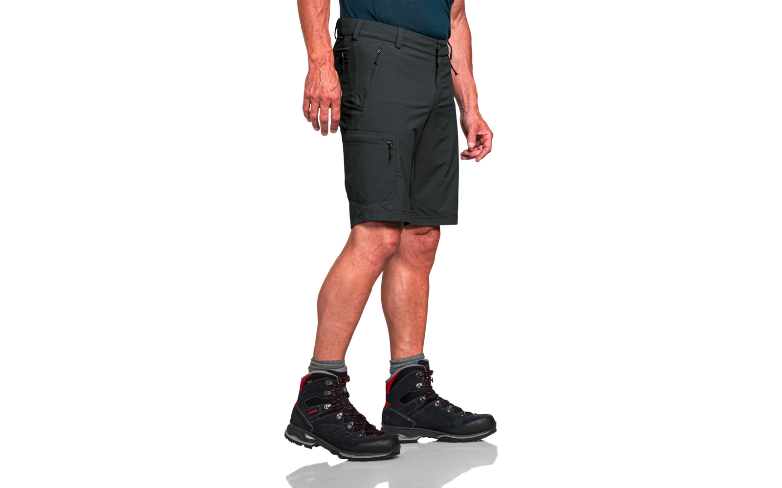 Unterkörper einer Person von der Taille bis zu den Füßen, dunkle knielange Shorts mit seitlicher Reißverschlusstasche, Hände an den Seiten, graue Knöchelsocken, schwarze Stiefel mit roten Details auf weißem Hintergrund