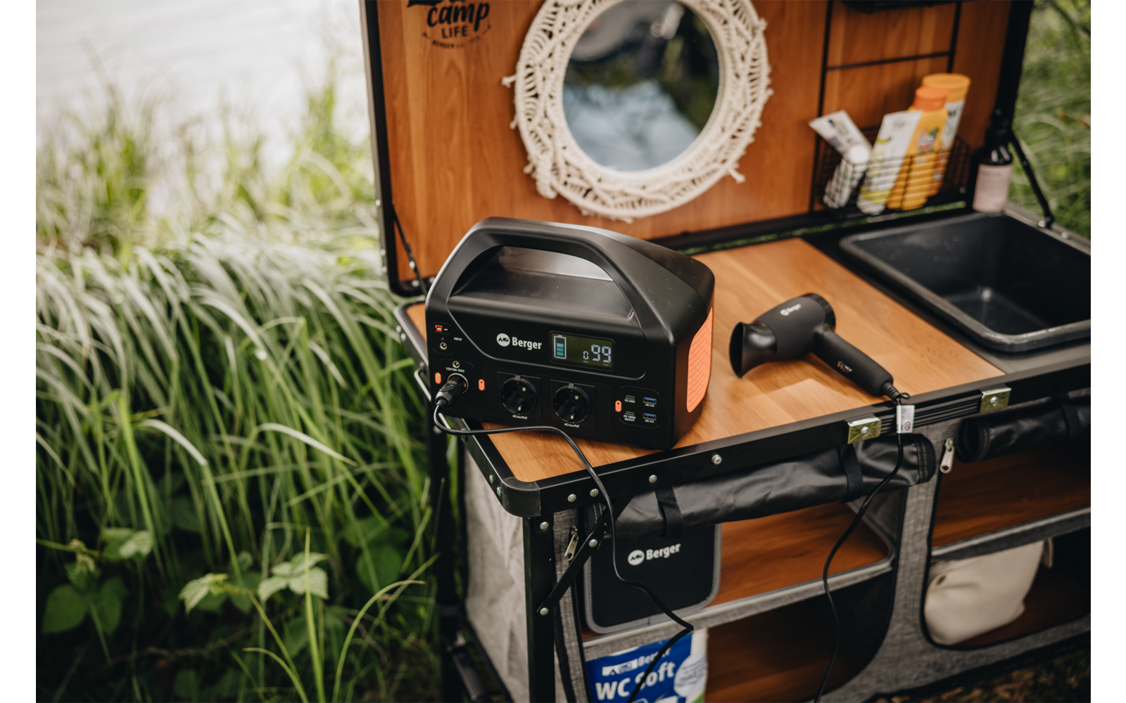 Station d'alimentation portable sur un plan de travail en bois avec un petit évier, un sèche-cheveux à main, un miroir rond, des brosses à dents et des hautes herbes au bord de l'eau