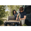 Femme aux cheveux foncés assise à une table de camping avec un ordinateur portable ; homme debout à côté d'elle manipulant un petit appareil ; cuisine pliante près d'un van ouvert, arbres en arrière‑plan