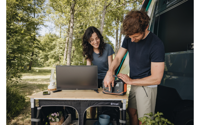 Femme aux cheveux foncés assise à une table de camping avec un ordinateur portable ; homme debout à côté d'elle manipulant un petit appareil ; cuisine pliante près d'un van ouvert, arbres en arrière‑plan