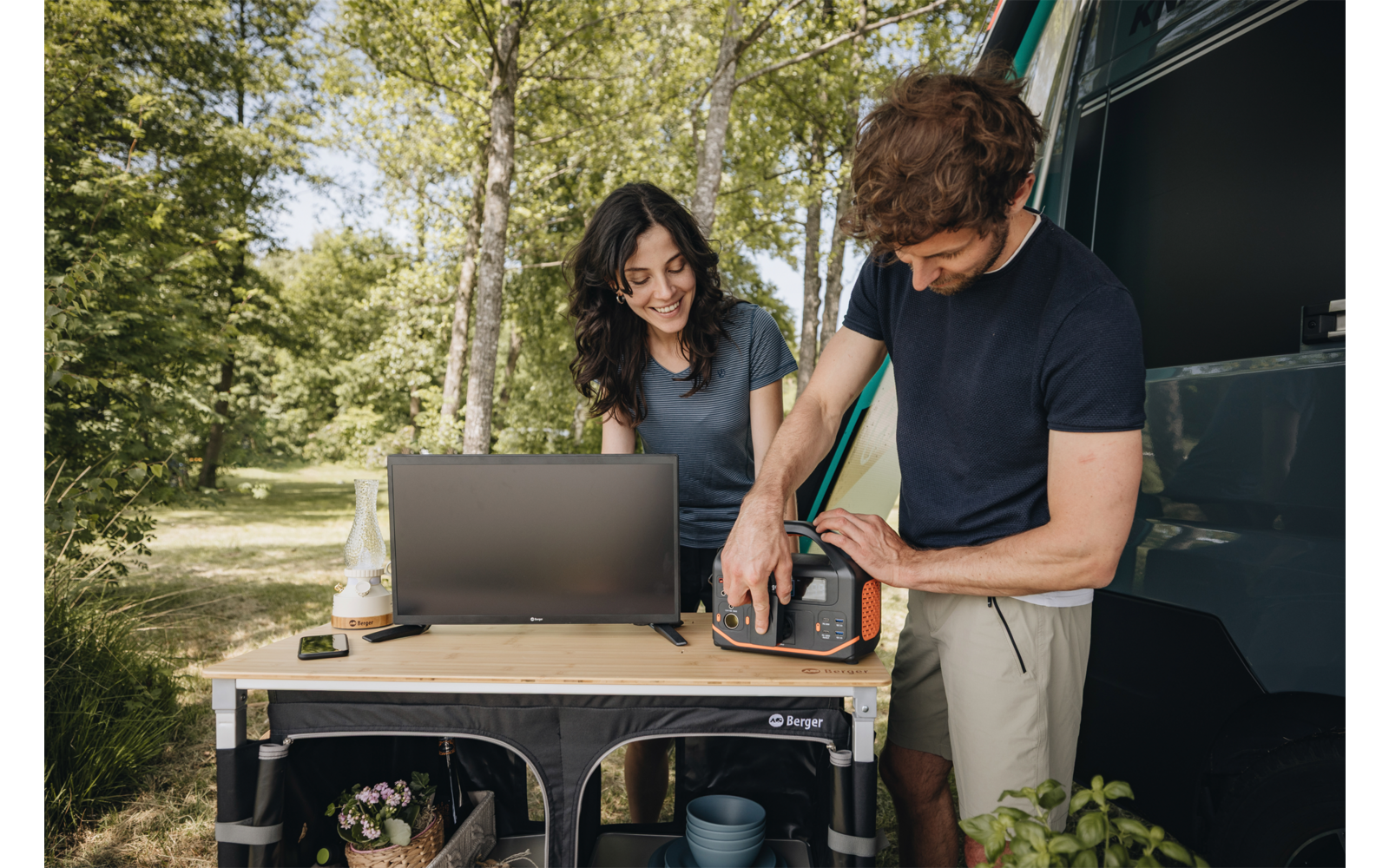 Femme aux cheveux foncés assise à une table de camping avec un ordinateur portable ; homme debout à côté d'elle manipulant un petit appareil ; cuisine pliante près d'un van ouvert, arbres en arrière‑plan