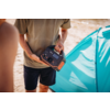 Buste d'une personne en t‑shirt vert olive et short foncé tenant une station d'alimentation portable noire et orange devant une tente turquoise sur du sable