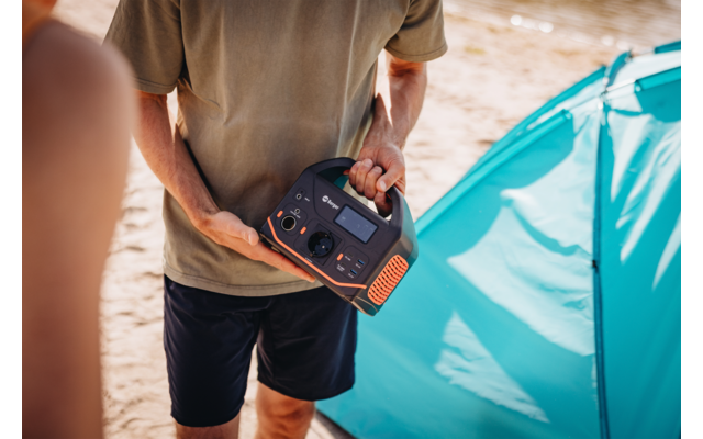 Buste d'une personne en t‑shirt vert olive et short foncé tenant une station d'alimentation portable noire et orange devant une tente turquoise sur du sable