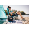 Personne portant des lunettes de soleil assise sur une chaise de camping sur une plage de sable, regardant un smartphone ; station d'alimentation portable sur une couverture au premier plan ; tente de plage turquoise et eau en arrière-plan