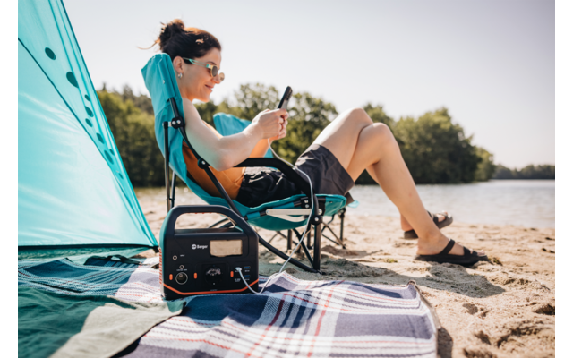 Personne portant des lunettes de soleil assise sur une chaise de camping sur une plage de sable, regardant un smartphone ; station d'alimentation portable sur une couverture au premier plan ; tente de plage turquoise et eau en arrière-plan