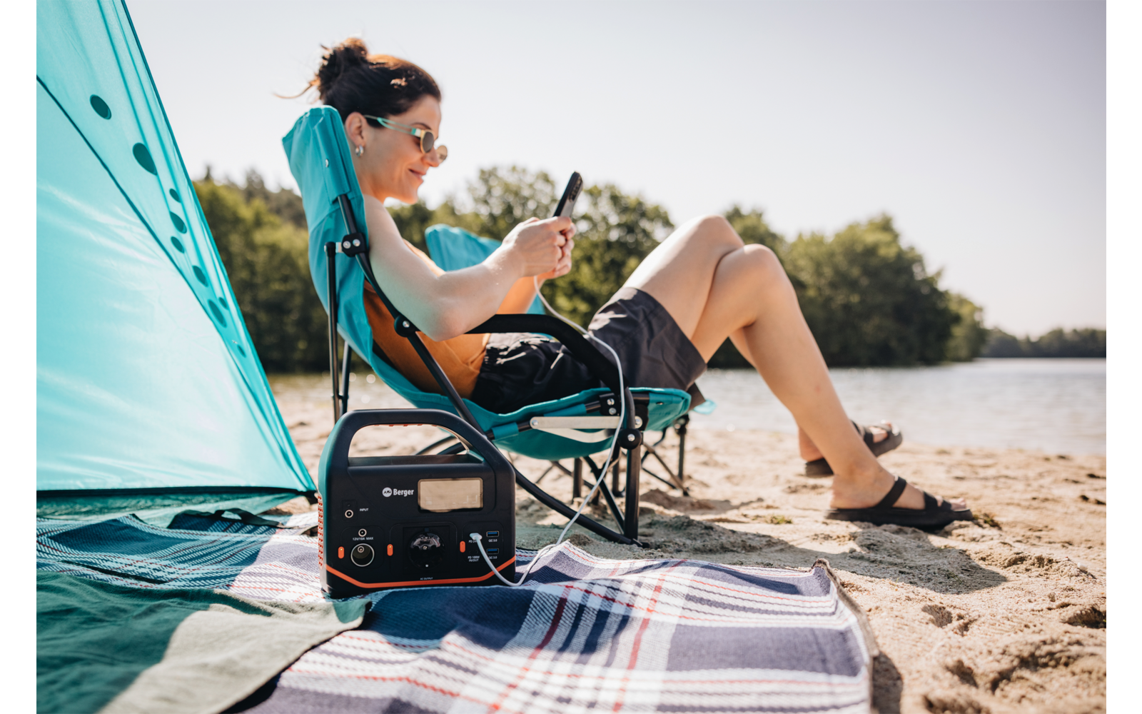 Personne portant des lunettes de soleil assise sur une chaise de camping sur une plage de sable, regardant un smartphone ; station d'alimentation portable sur une couverture au premier plan ; tente de plage turquoise et eau en arrière-plan