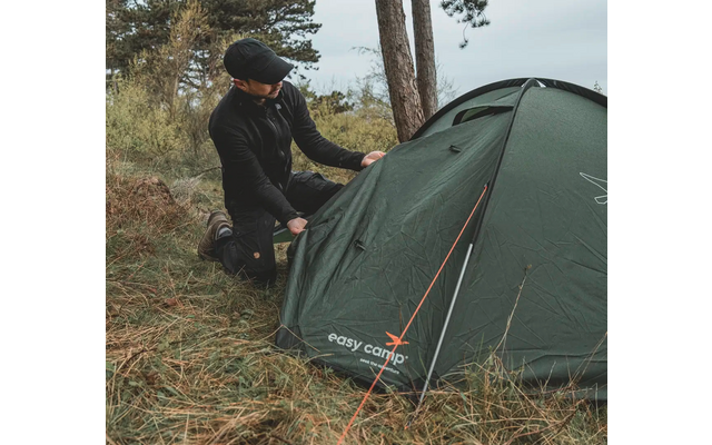 Person im Outdoor-Outfit neben grünem Zelt im Wald
