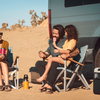 Deux personnes assises sur des chaises de camping, une personne assise sur les genoux de l'autre; une personne tient une glace rose; camionnette garée sur le sable, végétation désertique clairsemée en arrière-plan