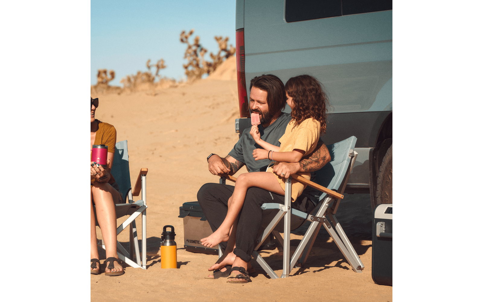 Deux personnes assises sur des chaises de camping, une personne assise sur les genoux de l'autre; une personne tient une glace rose; camionnette garée sur le sable, végétation désertique clairsemée en arrière-plan