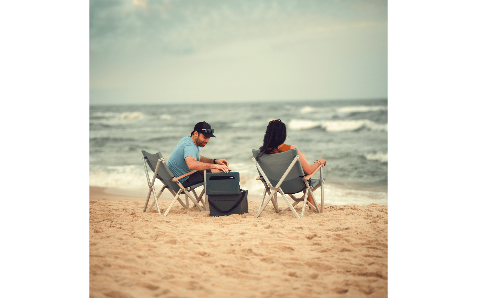 Deux personnes assises sur des chaises pliantes sur une plage de sable, une glacière entre elles, vagues et ciel nuageux en arrière-plan