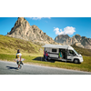 Camping-car blanc avec porte latérale ouverte et auvent déployé garé sur une bande herbeuse devant des montagnes rocheuses; une personne marche sur la route en portant un longboard, une autre personne se tient dans l'encadrement de la porte