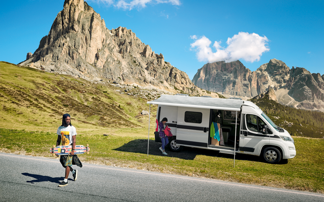Camping-car blanc avec porte latérale ouverte et auvent déployé garé sur une bande herbeuse devant des montagnes rocheuses; une personne marche sur la route en portant un longboard, une autre personne se tient dans l'encadrement de la porte