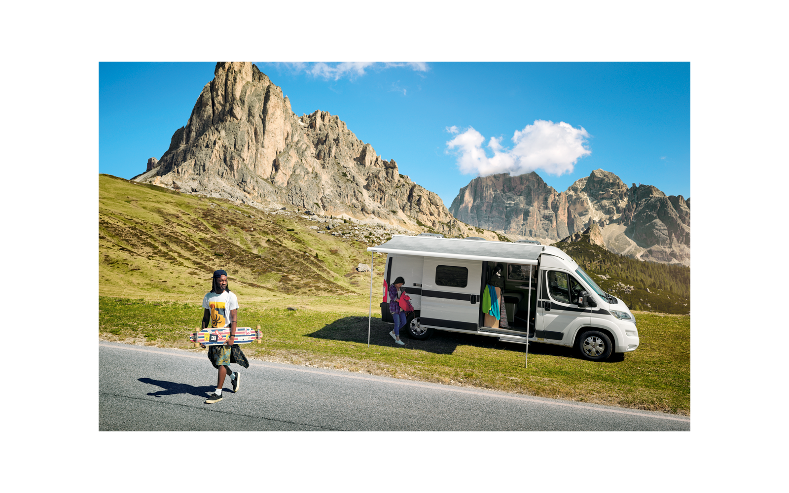 Camping-car blanc avec porte latérale ouverte et auvent déployé garé sur une bande herbeuse devant des montagnes rocheuses; une personne marche sur la route en portant un longboard, une autre personne se tient dans l'encadrement de la porte