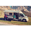 Camping-car blanc avec auvent déployé sur une prairie, une personne à côté et une autre dans la porte latérale ouverte, montagnes rocheuses en arrière-plan, route au premier plan