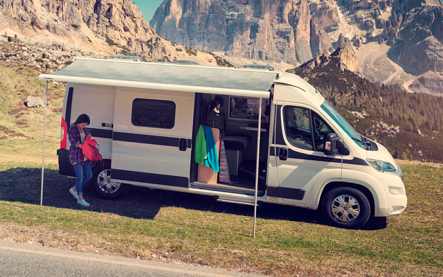 Camping-car blanc avec auvent déployé sur une prairie, une personne à côté et une autre dans la porte latérale ouverte, montagnes rocheuses en arrière-plan, route au premier plan