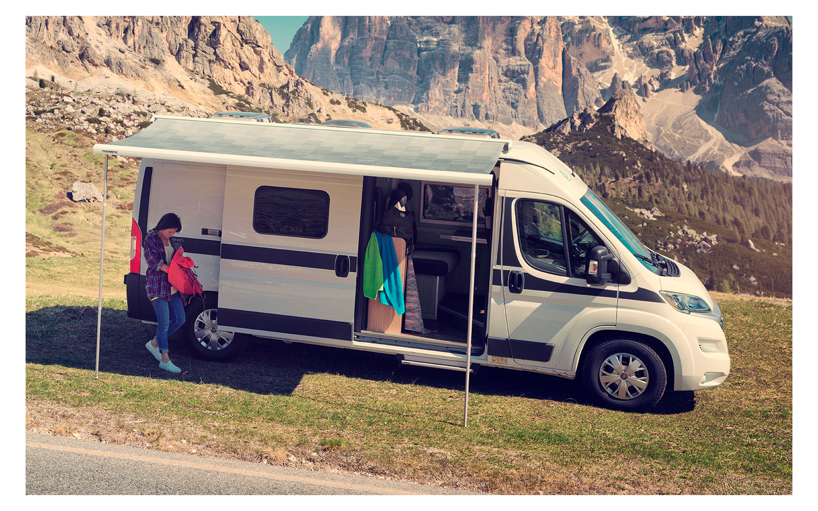 Camping-car blanc avec auvent déployé sur une prairie, une personne à côté et une autre dans la porte latérale ouverte, montagnes rocheuses en arrière-plan, route au premier plan