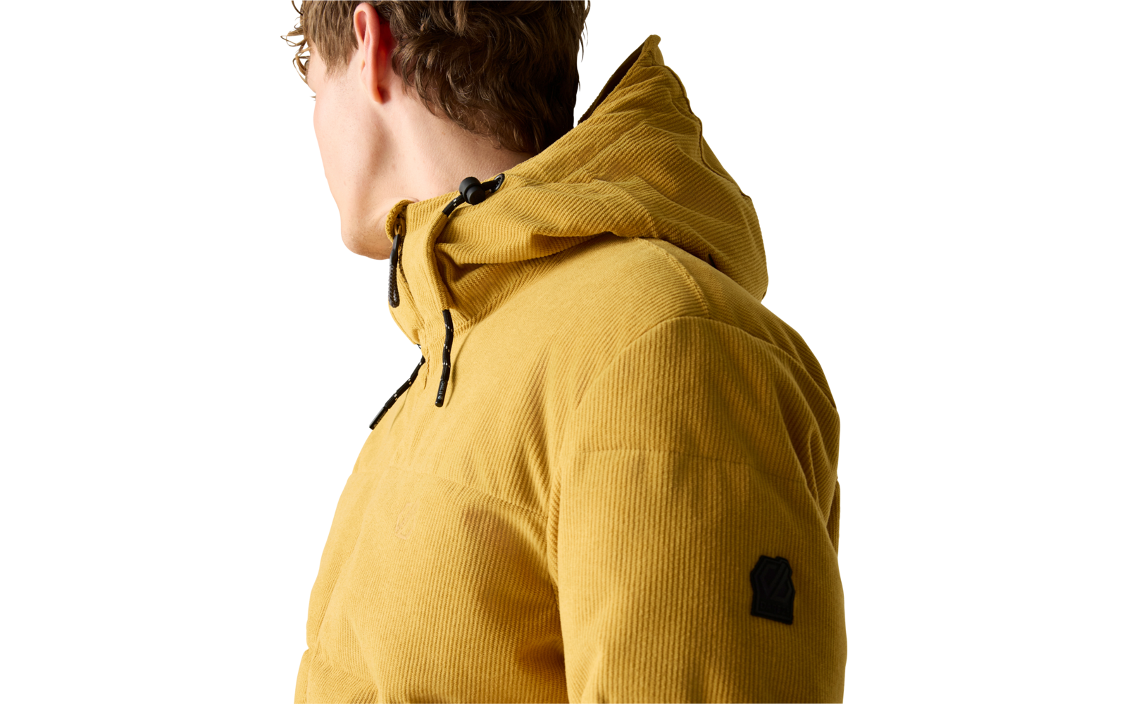 Person seitlich gesehen, trägt eine senfgelbe Cord‑Steppjacke mit Kapuze; sichtbar sind Kapuze, Reißverschlüsse, kleines schwarzes Patch an der Ärmelaußenseite und ein heller Hintergrund