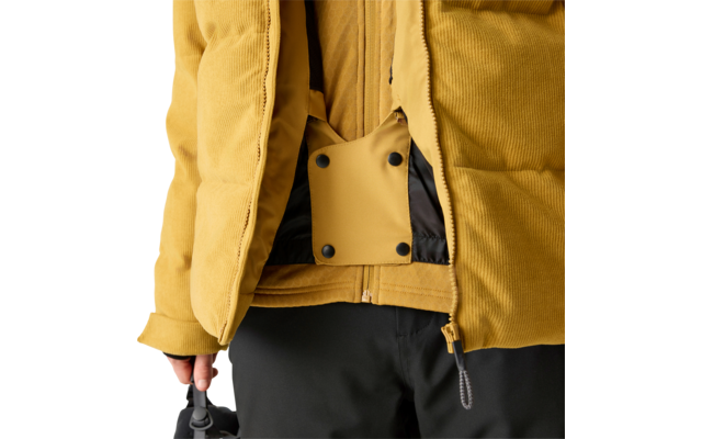 Oberkörper einer Person in senfgelber Cordjacke, Jacke geöffnet, innen sichtbares gelbes Riemenfeld mit vier schwarzen Druckknöpfen und Reißverschluss, schwarze Hose, linke Hand hält einen schwarzen Gegenstand, hellgrauer Hintergrund