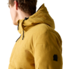Person seitlich gesehen, trägt eine senfgelbe Cord‑Steppjacke mit Kapuze; sichtbar sind Kapuze, Reißverschlüsse, kleines schwarzes Patch an der Ärmelaußenseite und ein heller Hintergrund