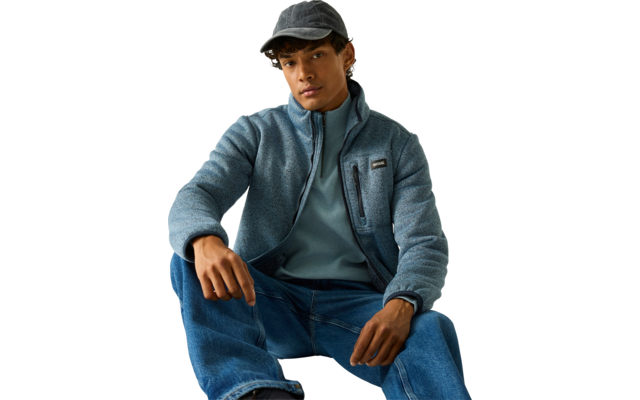 Zittend persoon met grijze pet, blauwe fleecejas met rits en borstzak, blauw sweatshirt eronder en spijkerbroek tegen een lichte achtergrond