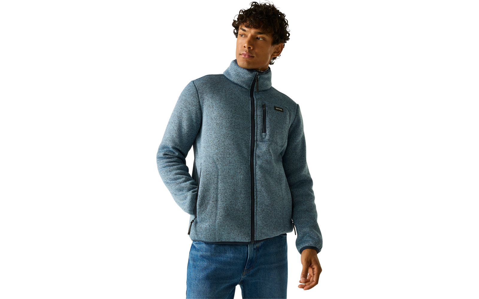 Man met krullend donker haar, grijsblauwe fleecejas met rits, handen in zakken, blauwe spijkerbroek, effen lichte achtergrond, kijkt naar rechts