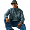 Zittend persoon met grijze pet, blauwe fleecejas met rits en borstzak, blauw sweatshirt eronder en spijkerbroek tegen een lichte achtergrond