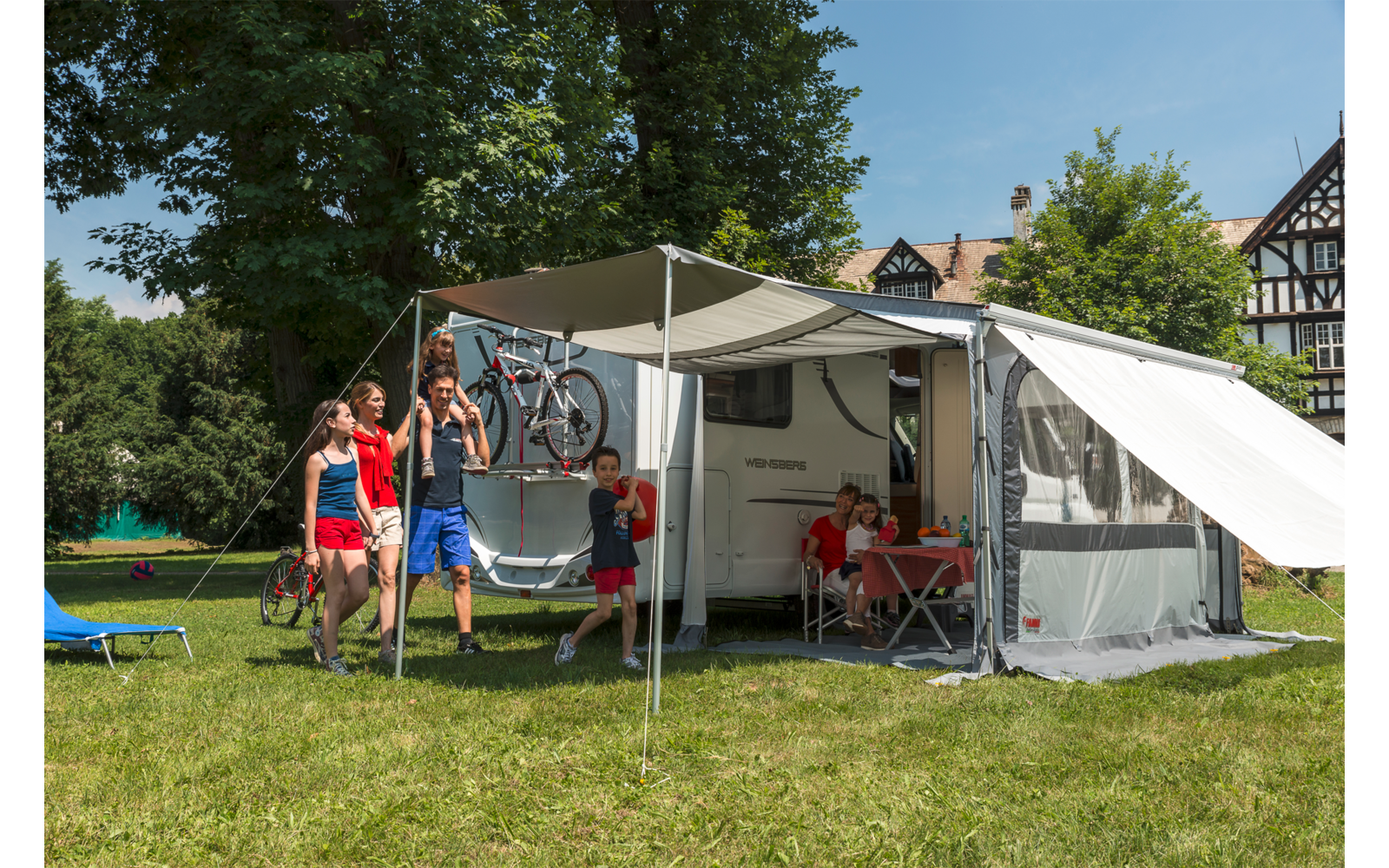 Wohnwagen mit Markise auf einer Wiese, davor zwei Erwachsene und zwei Kinder, Fahrrad am Wohnwagen, Tisch und Stühle unter der Markise, Fachwerkhäuser, Bäume und blauer Himmel