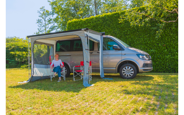 Silberner Campervan mit ausgefahrener Markise; Frau sitzt auf einem Campingklappstuhl darunter; roter Klappstuhl und kleiner Tisch neben ihr; Hecke und Bäume im Hintergrund; Rasen im Vordergrund
