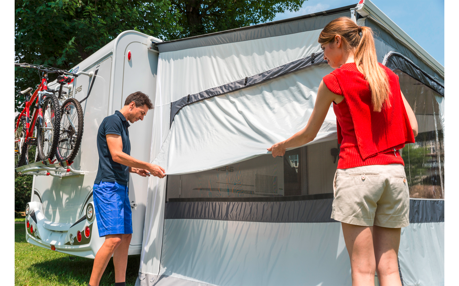 Zwei Personen, Mann in dunklem T-Shirt und blauen Shorts und Frau in rotem Top und beigen Shorts halten ein weißes Textil mit Sichtfenster, das an einem weißen Wohnwagen befestigt ist; zwei Fahrräder am Heck; Gras und Bäume im Hintergrund