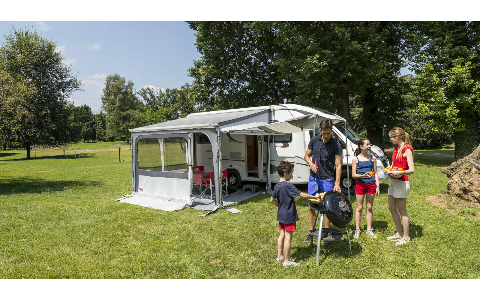 Autocaravana con toldo desplegado en un césped; cuatro personas de pie junto a una barbacoa de forma redonda; árboles y sillas de campaña al fondo